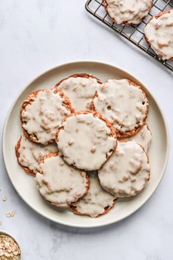 Iced oatmeal cookies on a plate with wire rack of more cookies nearby.