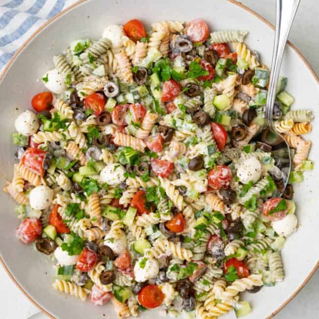 Italian pasta salad in a large serving bowl with a spoon dipped in a serving plates nearby.
