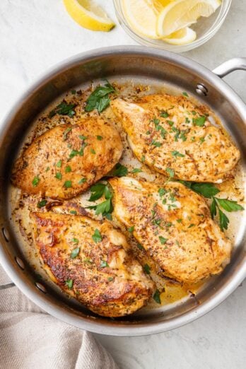 Juicy pan seared chicken breast in a stainless teel pan garnished with fresh parsley with a small bowl of lemon wedges nearby.