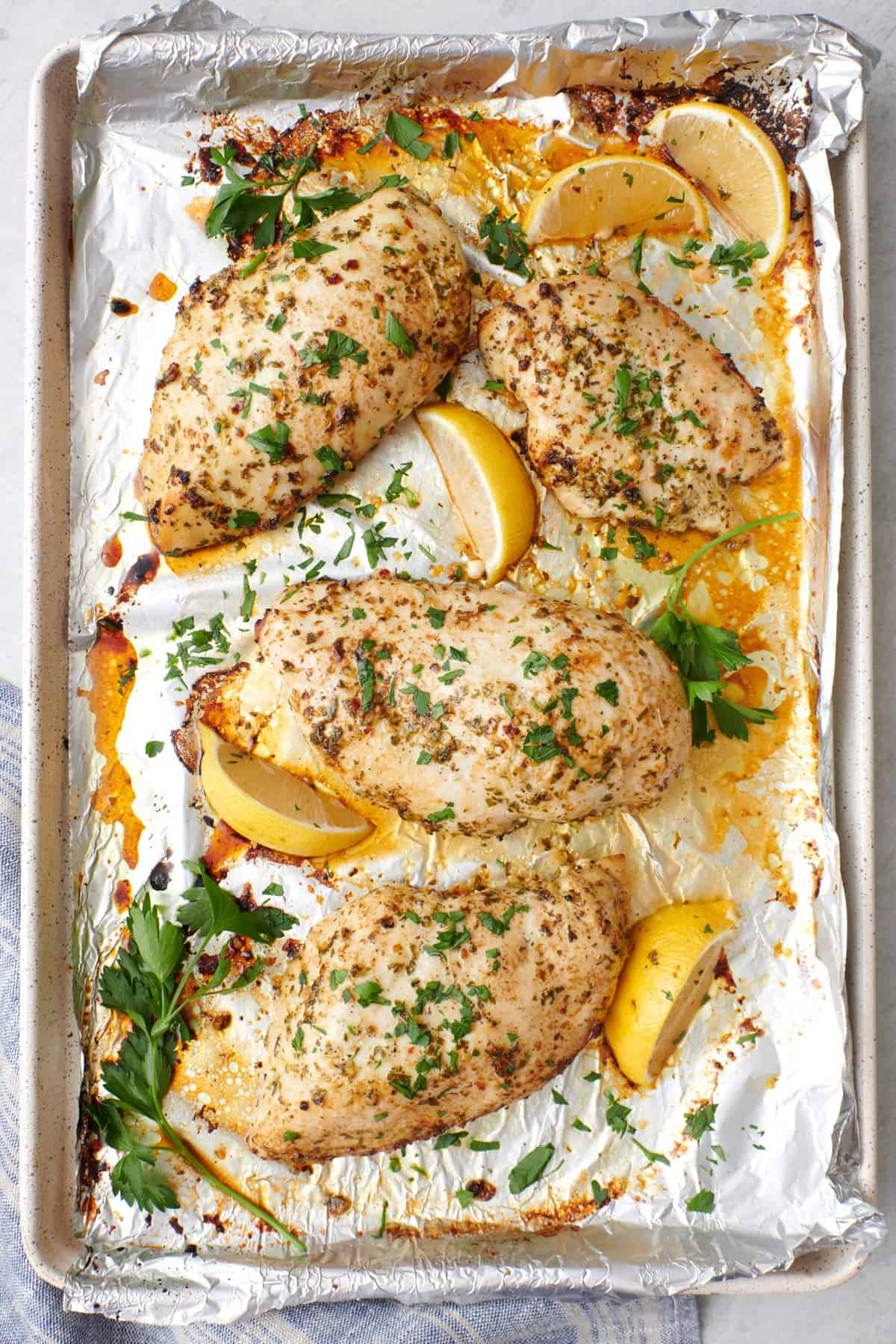 Sheet pan lemon garlic chicken breasts on a foil lined cooking sheet, garnished with fresh parsley and lemon wedges.