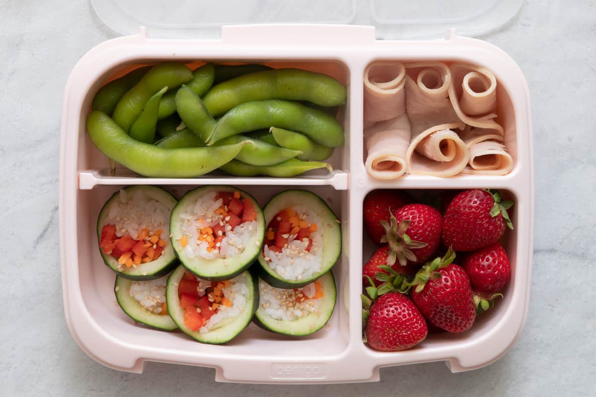 Lunchbox with 4 sections with different foods in each section: edamame in shell, turkey roll ups, cucumber sushi, and whole fresh strawberries.