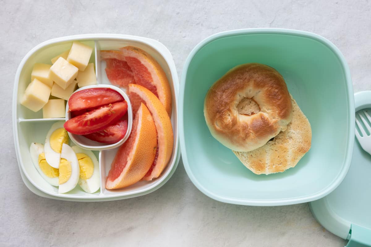 Stackable lunch container with individual sections and different foods in each: large container filled with toasted bagel, sectioned container has cheese cubes, quartered boiled eggs, tomato wedges, and slices of grapefruit.