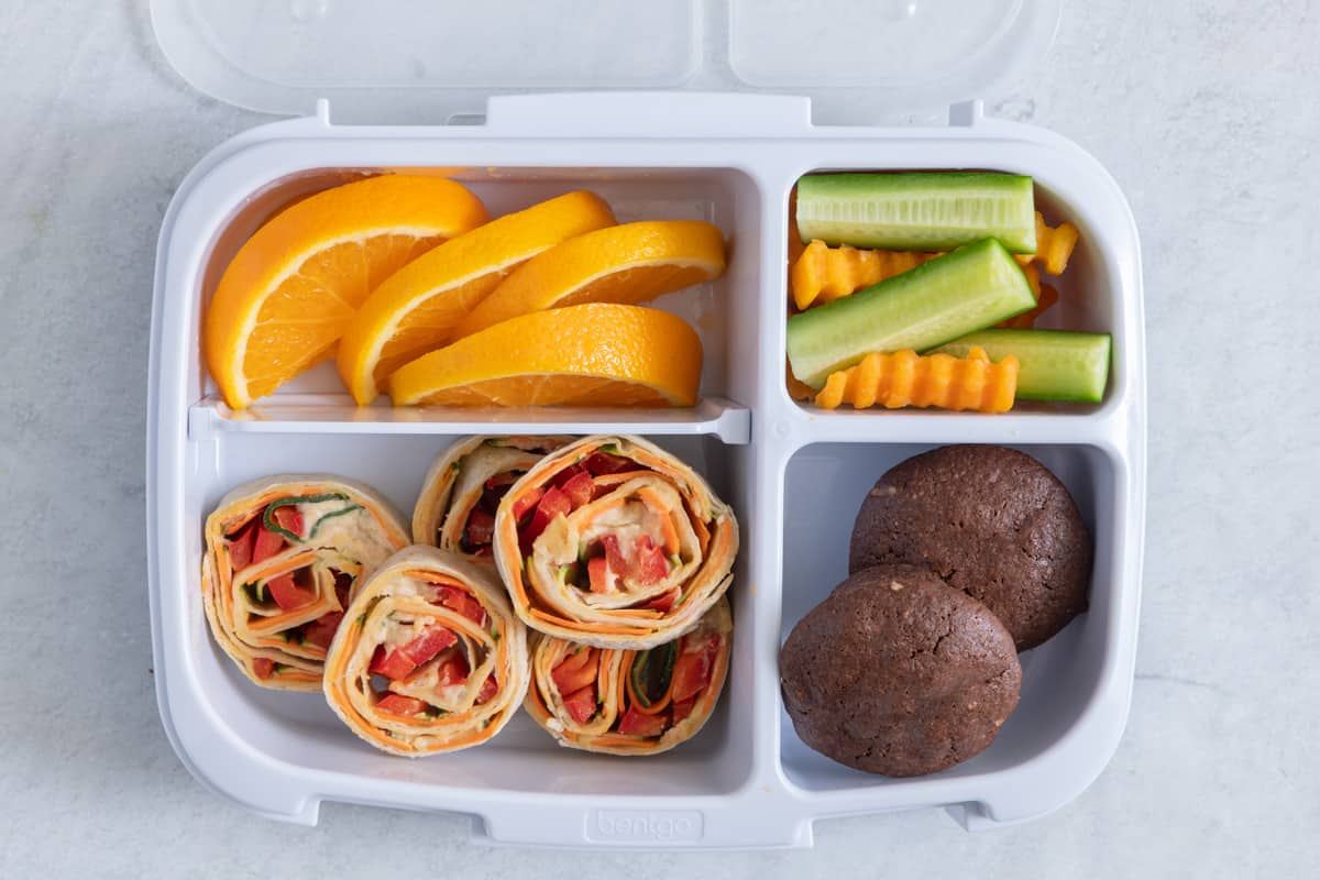 Lunchbox with 4 sections with different foods in each section: sliced oranges, crinkle carrots and cucumbers, 2 mini chocolate muffins, and 5 hummus veggie pinwheels.