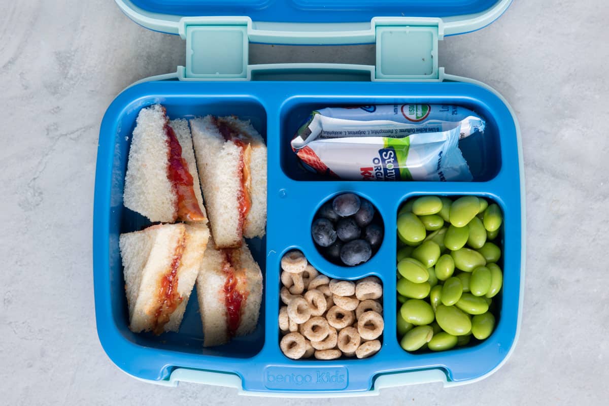 Lunchbox with individual sections with different lunch foods for kids: peanut butter and jelly sandwich cut into triangles, Stonyfield yogurt tube, blueberries, Cherrioes, and edamame.