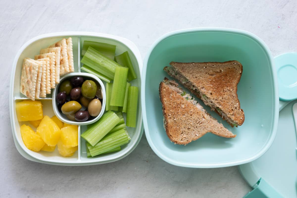 Stackable lunch container with individual sections and different foods in each: saltine crackers, pineapple chunks, olives, and celery sticks with a tuna sandwich in a large container.