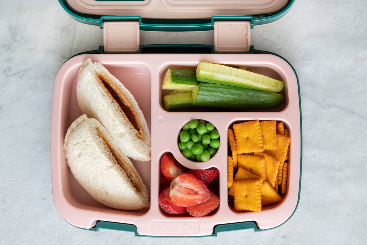 Lunchbox with individual sections with different lunch foods for kids: Uncrustable sandwich cut in half, cucumber sticks, green peas, halved strawberries, and cheddar square crackers.