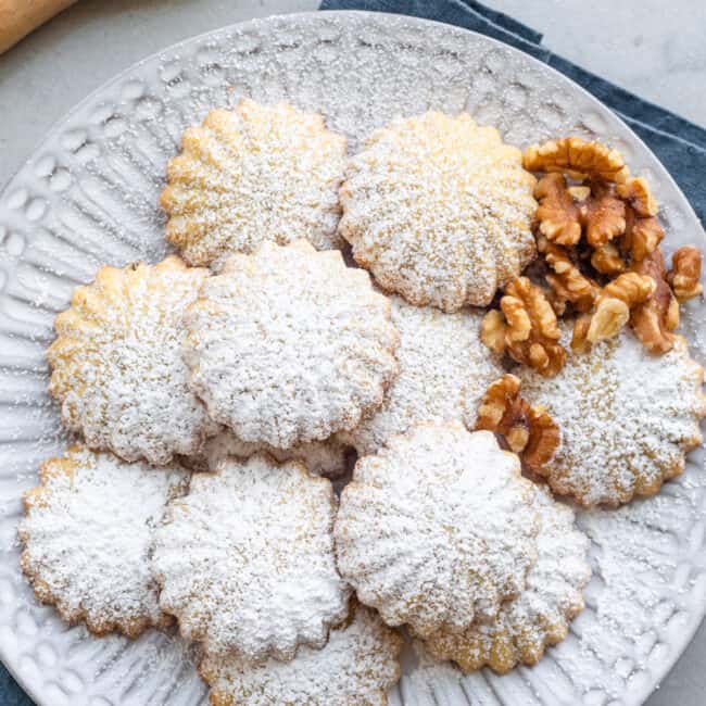 Maamoul cookies on a round decorative plate with extra walnuts dusted with powdered sugar.