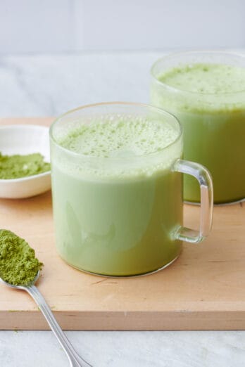 Matcha latte in a glass mug with matcha powder on a spoon and small dish.