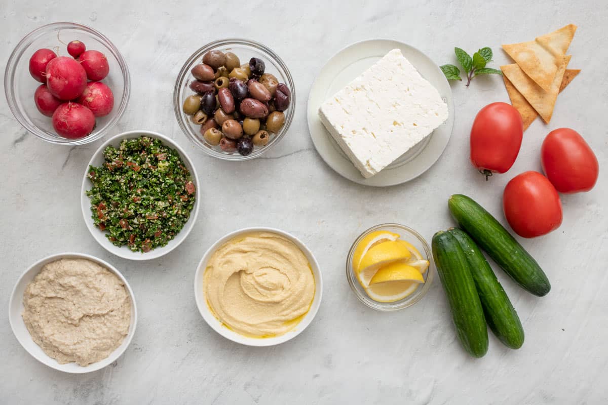 Ingredients to make board: radishes, tabbouleh, baba ghanoush, hummus, mixed olives, feta, lemon wedges, pita chips, mint leaves, tomatoes, and Persian cucumbers.