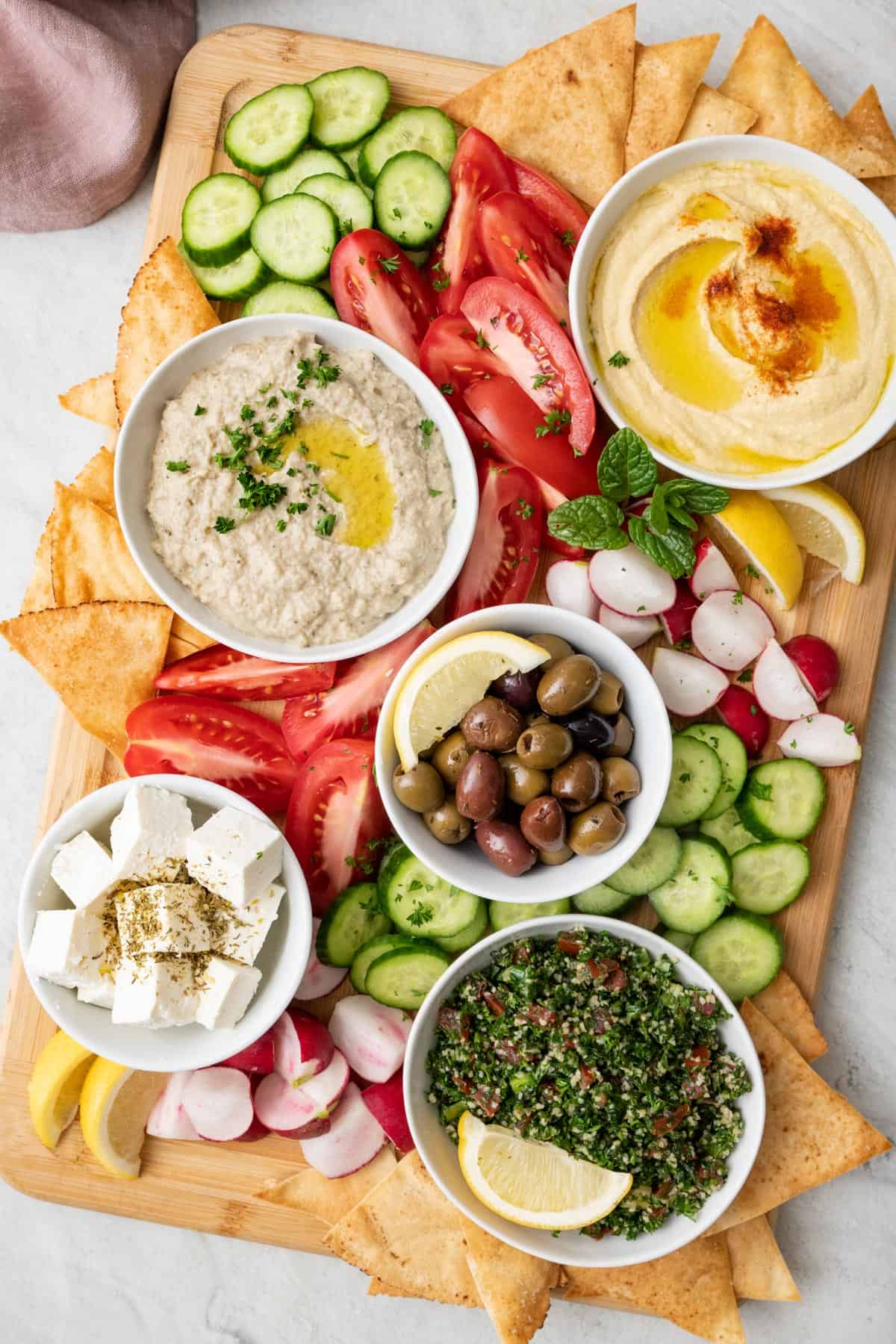 Final mezze platter with small bowls of hummus, baba ghanoush, small blocks of feta, olives, and tabbouleh, with quartered tomatoes and radishes, and sliced cucumbers spread in between bowls. Pita triangles tucked into the edges of the board and lemon garnished throughout.