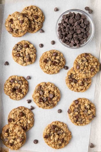 Baking sheet lined with parchment paper with baked cookies layered on top of eachother and a small dish of chocolate chips in the corn.