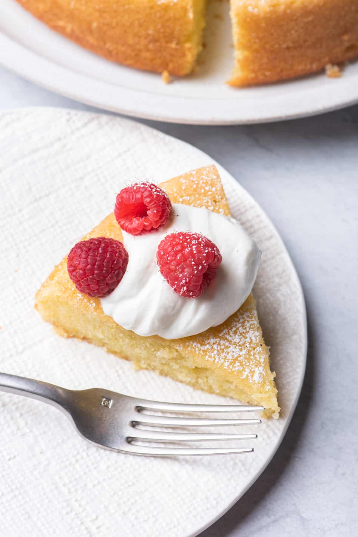Slice of olive oil cake on a small plate topped with whipped cream and raspberries