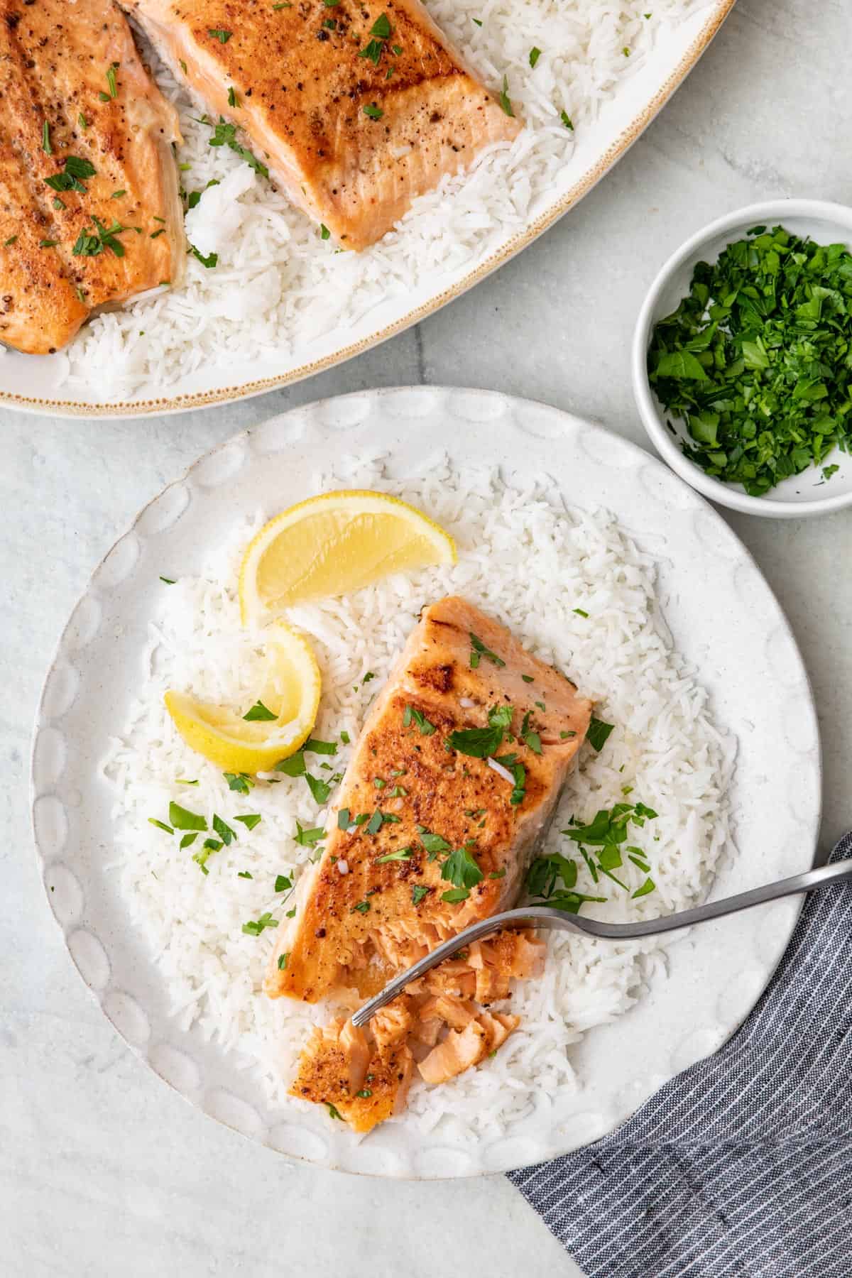 One salmon served over rice on a small serving plate with platter nearby ganished with lemon and parsley.