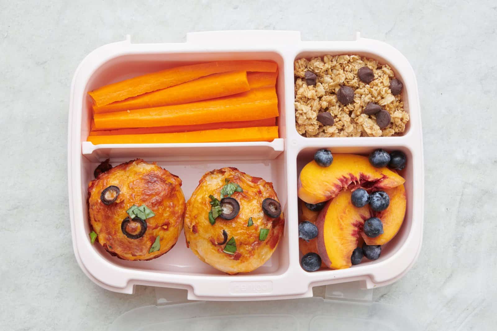 Pizza muffin lunch box with carrot sticks, granola, and sliced peaches and blueberries.