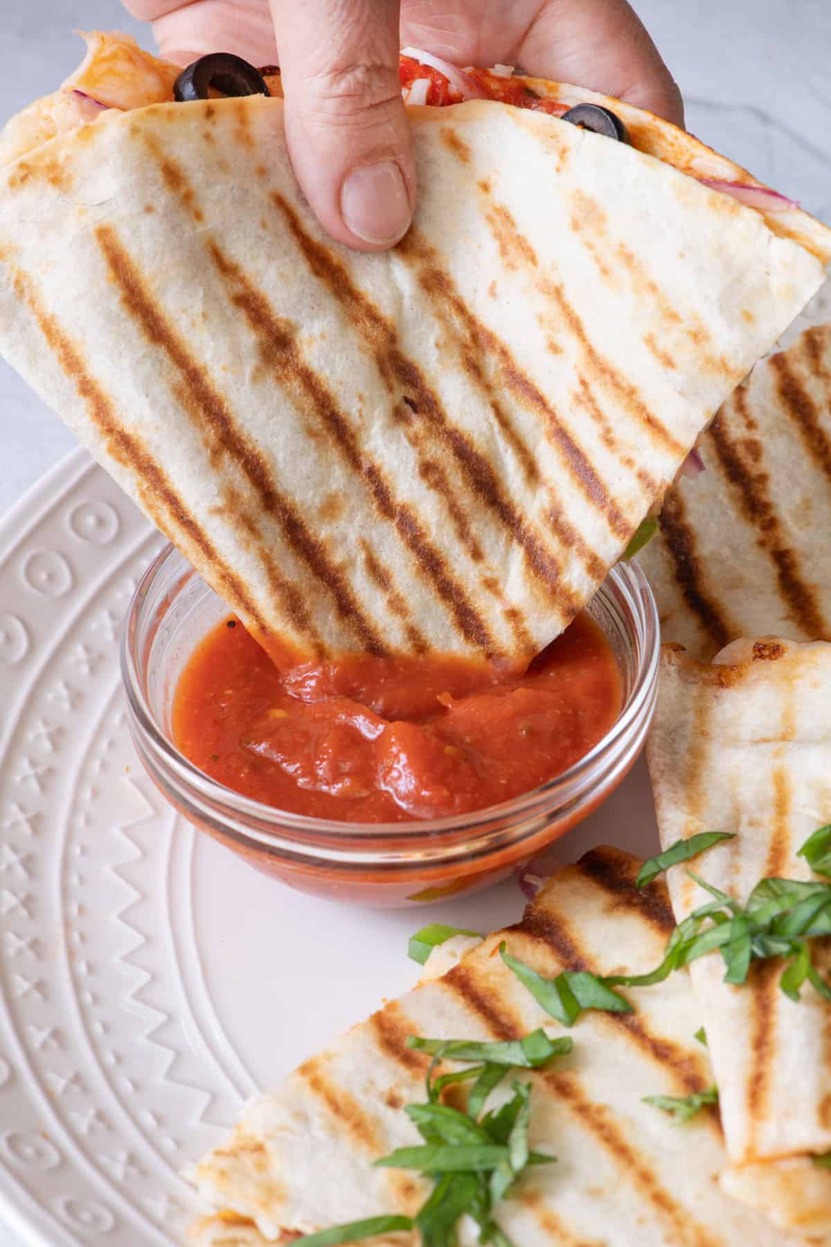 A pizzadilla half being dipped into marinara sauce on a plate with more slices.