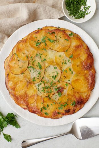Potato galette on a serving plate with a spatula nearby, garnished with fresh chopped parsley.