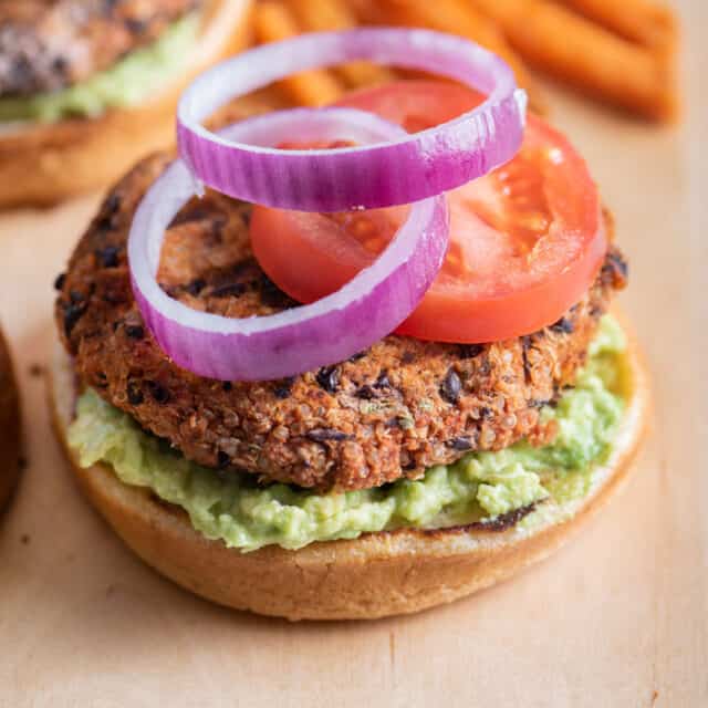 One quinoa burger topped with tomatoes and onions with sweet potato fries in background