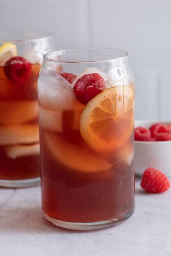 Two tall glasses of Raspberry Iced Tea lemon slices and fresh raspberries and a small white dish of raspberries.
