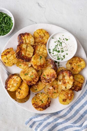Roasted parmesan potatoes on a serving dish cut side up to show crosshatch and crispy parmesan with a small pinch bowl of ranch, garnished with fresh parsley and serving spoon sitting on plate.
