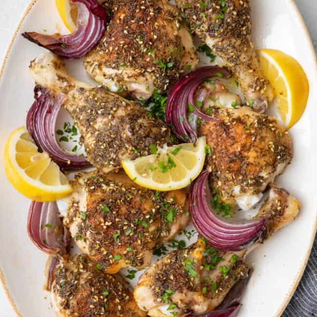 Roasted Zaatar chicken arranged on an oval platter with seasoned skin, garnished with red onion slices, lemon wedges, and fresh parsley.