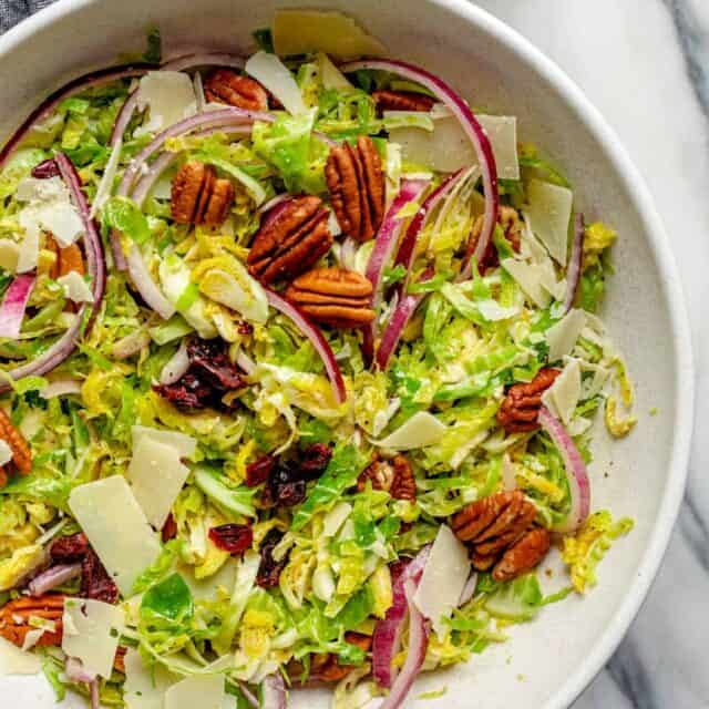 Large bowl of shaved brussel sprouts salad topped with pecans, onions, cheese, cranberries and red onions