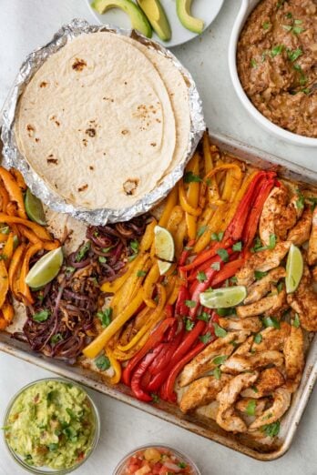 Sheet pan chicken fajitas garnished with fresh cilantro with small dishes of guacamole, salsa, flour taco shells, refried beans and sliced avocados.
