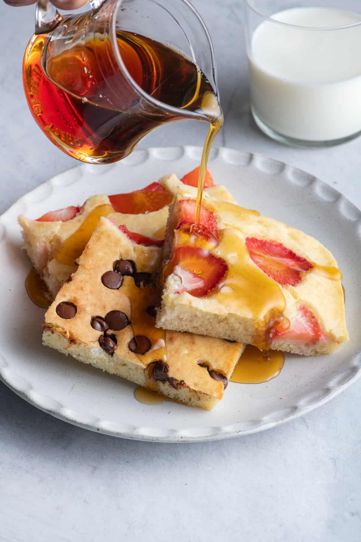 Syrup being poured on top of a stack of different flavor sheet pan pancakes on a white plate with a glass of milk nearby.