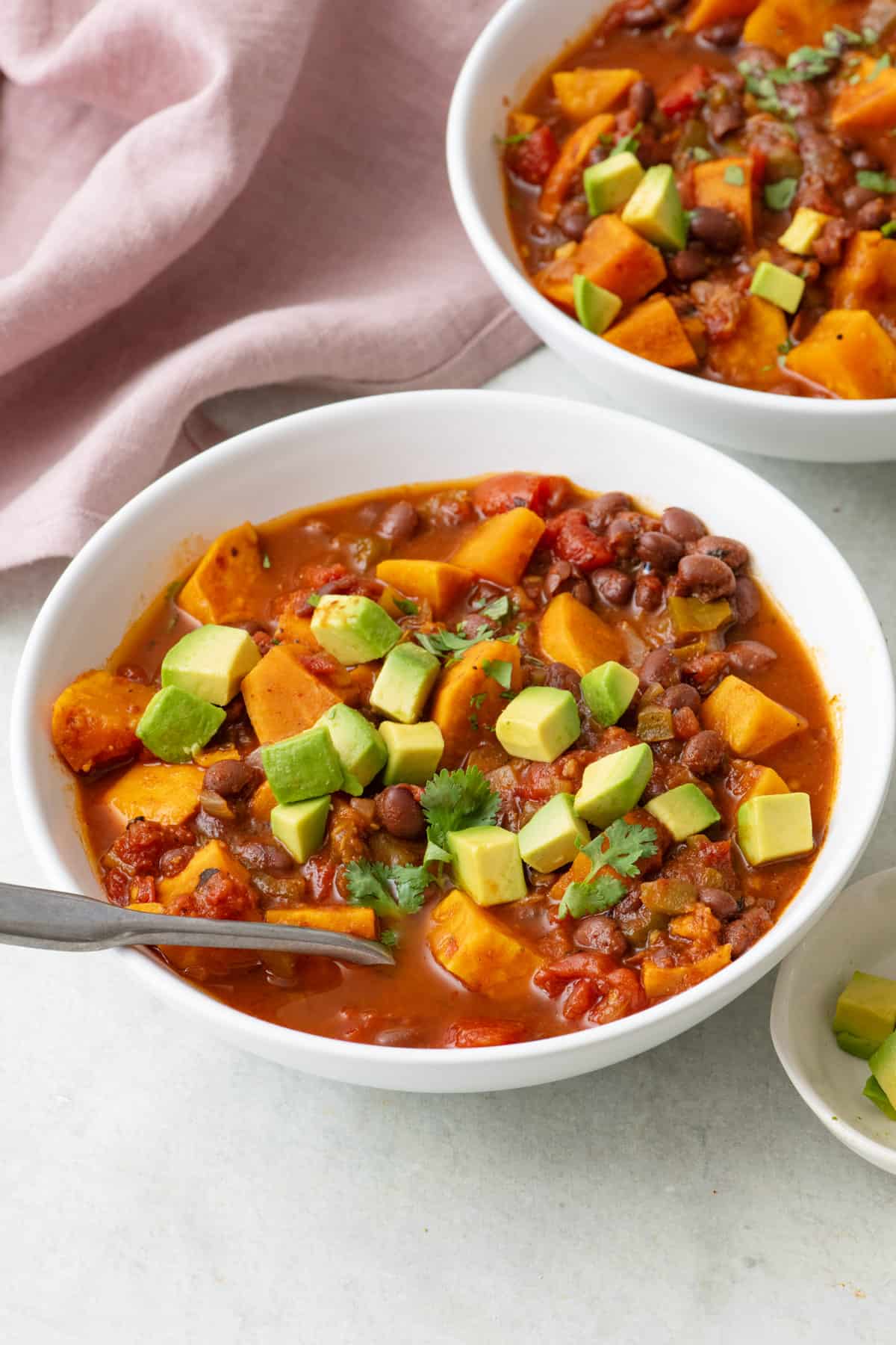 Bowl of sweet potato chili topped with diced avocado and a spoon dipped in. Another bowl of chili nearby.