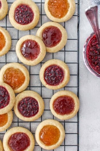 Three different jam filled thumbprint cookies on a wire rack with a jar of jam nearby.