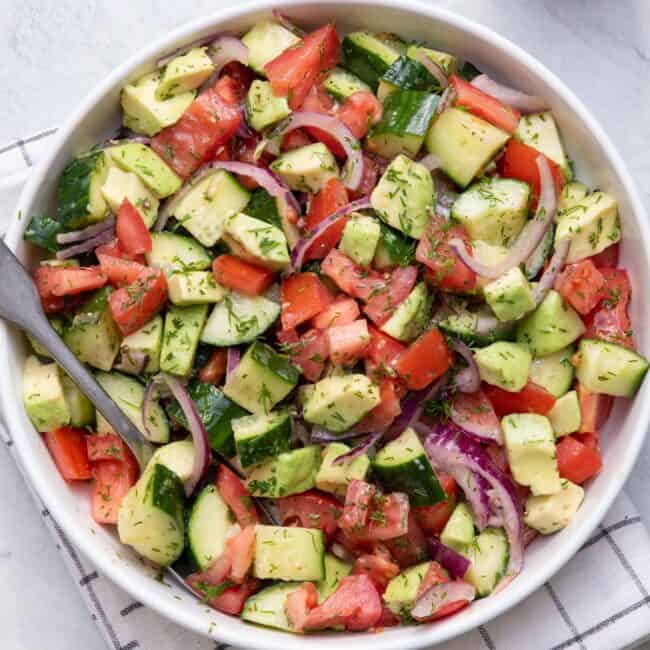 Big white bowl of Tomato Avocado Cucumber Salad all mixed up with a spoon in the large bowl.