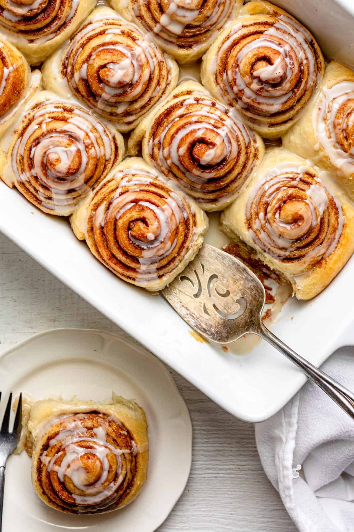 Large pan of 12 cinnamon rolls at an angle and one roll served on small dessert plate nearby