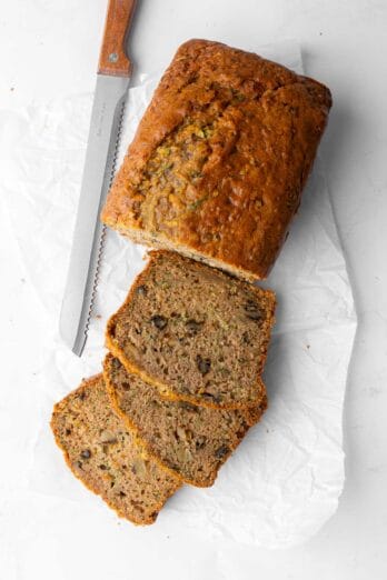 Zucchini bread on parchment paper cut halfway up into thick slices with a serrated knife nearby.